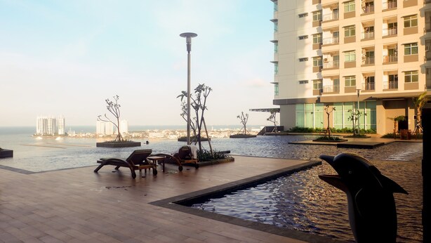 A rooftop pool area with lounge chairs, a dolphin sculpture, and a view of the sea and buildings. - Warm And Homey 2Br Apartment At Green Bay Pluit (Jakarta)