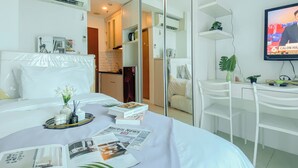 Habitación de hotel con cama, escritorio, silla, televisión y cocina visible a través de una puerta de vidrio.