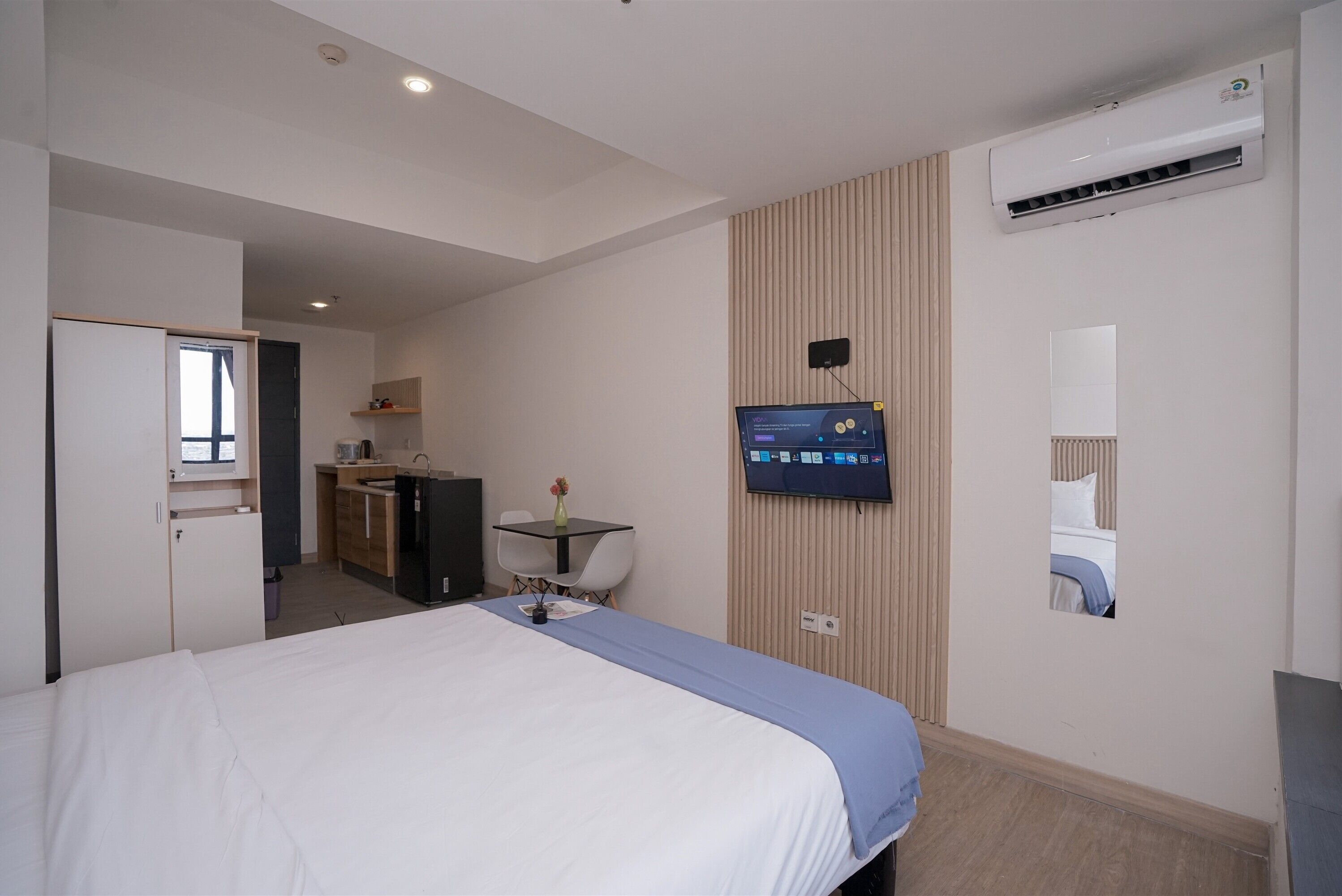 A hotel room with a bed, a desk with a computer, a small table with a vase, a wall-mounted TV, and an air conditioning unit.