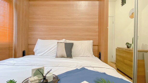 Un dormitorio con cabecera de madera, una cama con ropa de cama blanca, una mesita de noche con una planta y una ventana con persianas.