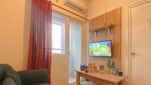 Una sala de estar con un sofá, un televisor sobre un soporte de madera, una mesita con libros y una planta, y una ventana con cortinas rojas.
