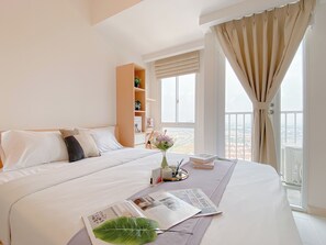 A hotel room with a large bed, a bedside table with a book and a vase of flowers, a window with a view, and a wooden shelf with decorative items. - Cozy And Enjoy Studio At Tokyo Riverside Pik 2 Apartment (Teluknaga)