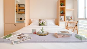 A neatly arranged hotel room with a bed, a side table with a magazine, a tray with a vase and candles, a bookshelf with books and a plant, and a window with a view. - Cozy And Enjoy Studio At Tokyo Riverside Pik 2 Apartment (Teluknaga)