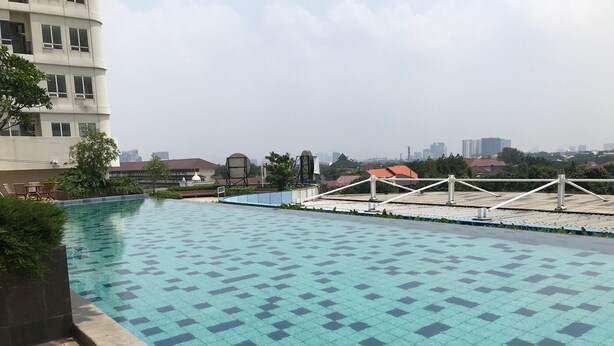 A rooftop swimming pool with a tiled floor, surrounded by greenery and a view of buildings in the distance. - Warm Stay Studio At Cinere Bellevue Suites Apartment (Depok)