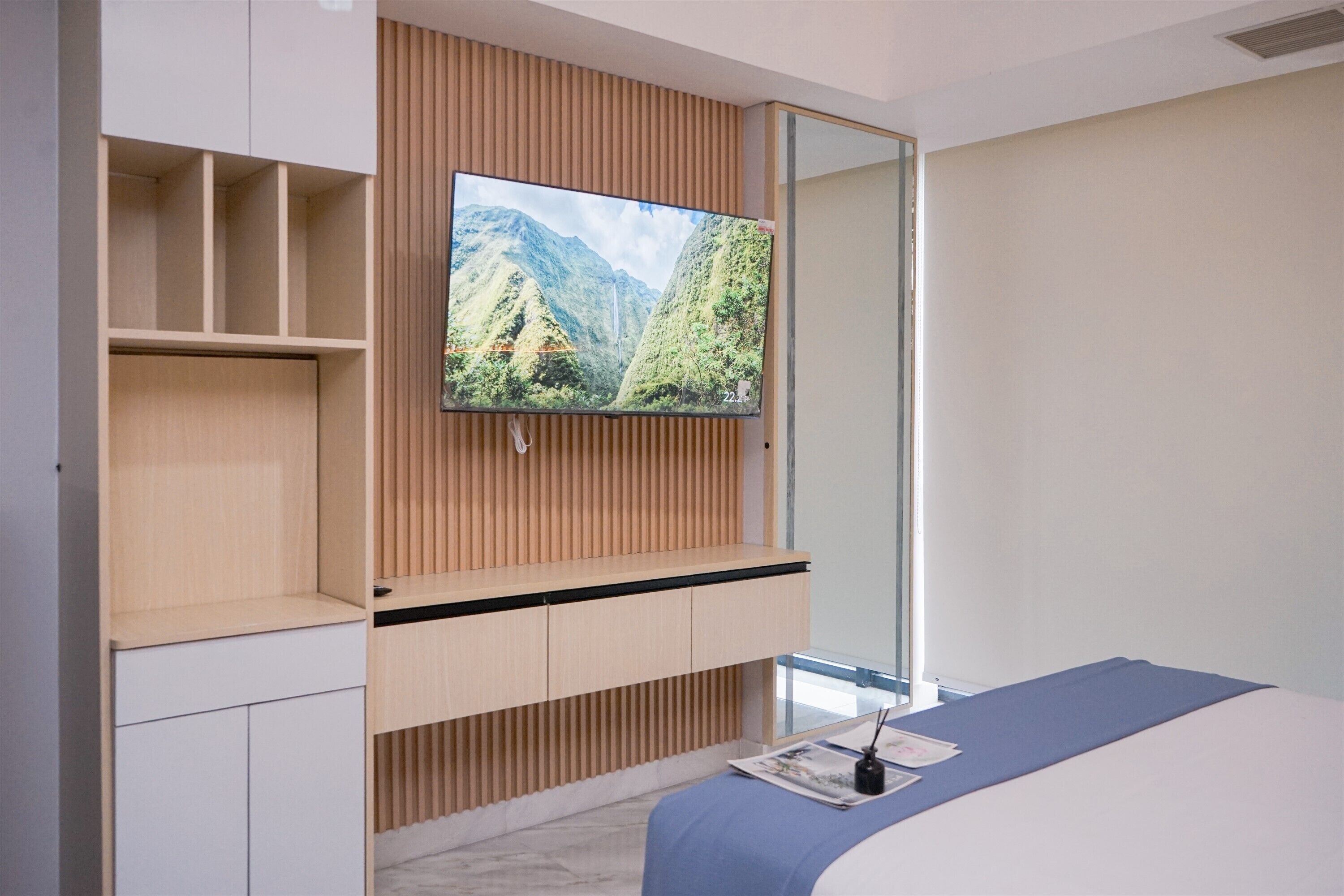 Una moderna camera d'albergo con una grande televisione a schermo piatto montata su un pannello di legno, un letto con copriletto blu e un tavolino con una rivista e una bottiglia.