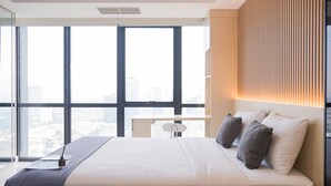 A modern hotel room with a large bed, a desk, and a view of the city through large windows. - Spacious Studio At Apartment The Smith Alam Sutera (Tangerang)