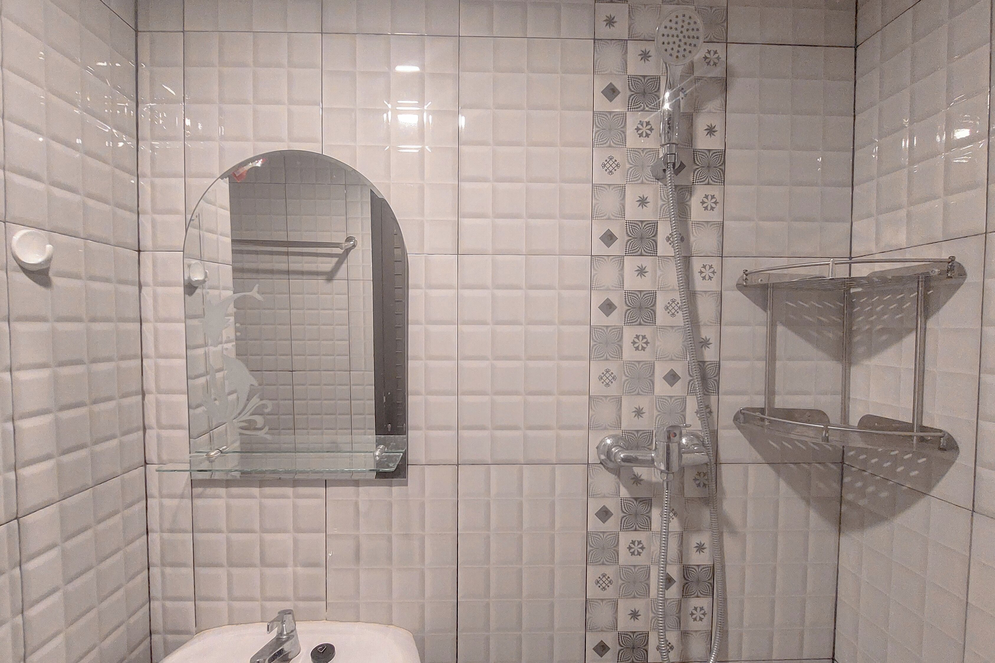 Un bany amb parets revestides de rajoles blanques, un cubicle de dutxa de vidre, un lavabo amb aixeta i un mirall sobre el lavabo.
