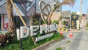 A sign for De'Prima Apartments with a heart-shaped logo, surrounded by greenery and a palm tree.