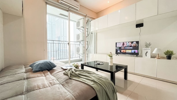A modern living room with a sofa, a coffee table, a TV, and a balcony with a city view. - Nice And Cozy 3Br Apartment At Thamrin Residence (Jakarta)
