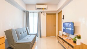 A modern living room with a grey sofa, a wooden TV stand with a flat-screen TV, a wooden cabinet, and a door leading to another room. - Comfy Stay 2Br At Meikarta Apartment (Cikarang)