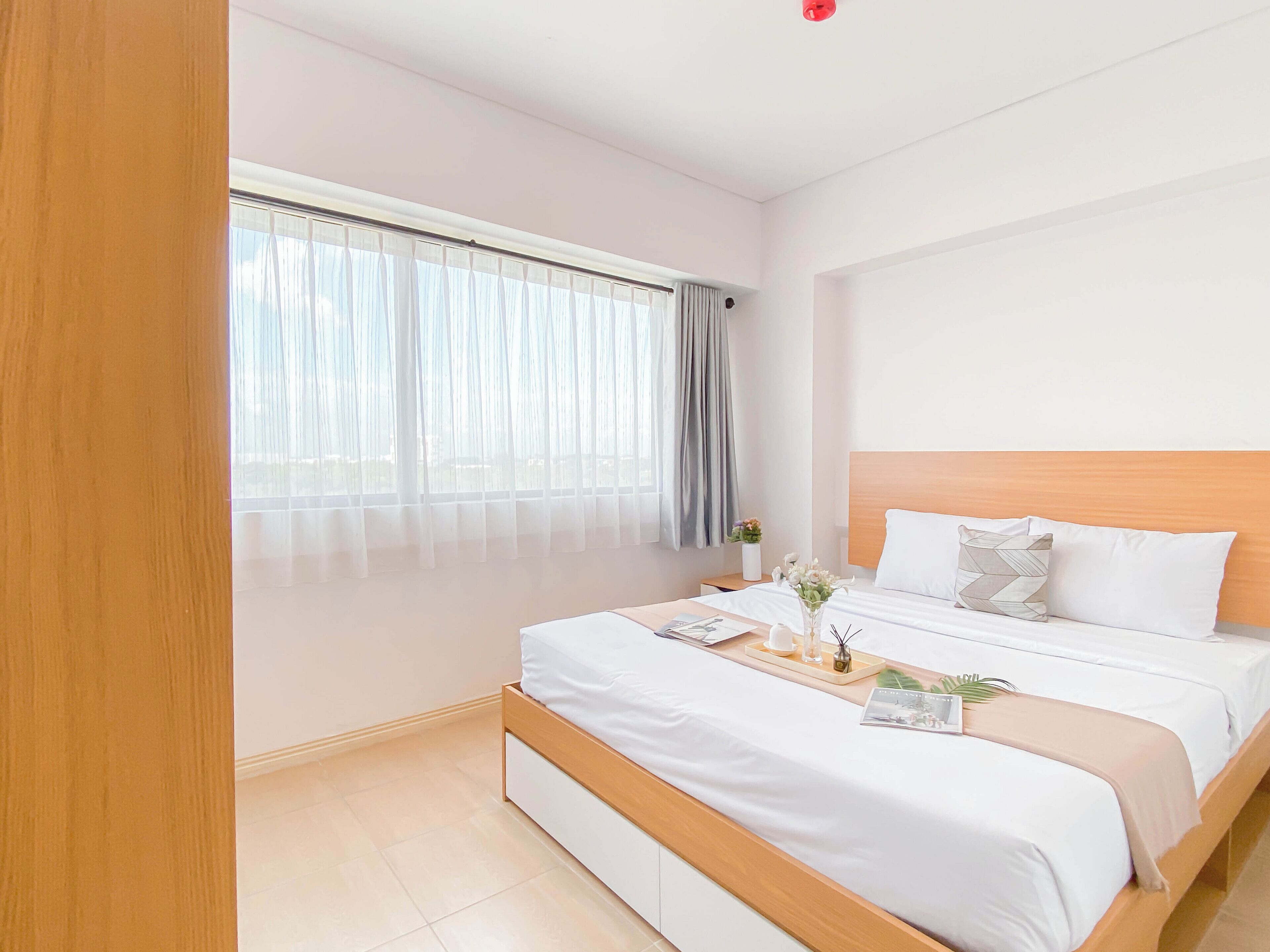 A modern hotel room with a large bed, a bedside table with a vase of flowers, and a window with sheer curtains.
