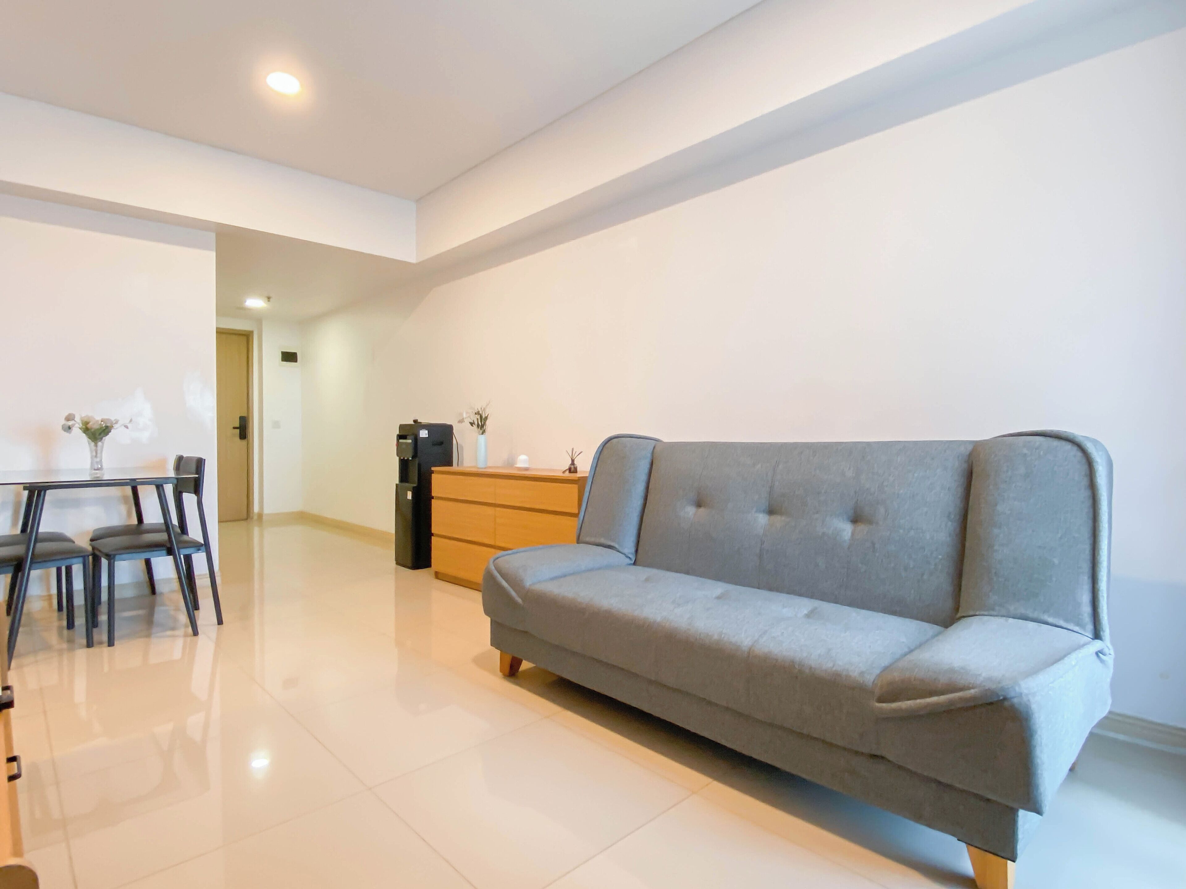 A modern living room with a grey sofa, a wooden dresser, a coffee machine, and a dining table with chairs.