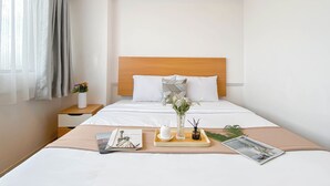 A neatly arranged hotel room with a bed, a bedside table with a vase of flowers, and a tray with a cup and a book. - Comfy Stay 2Br At Meikarta Apartment (Cikarang)
