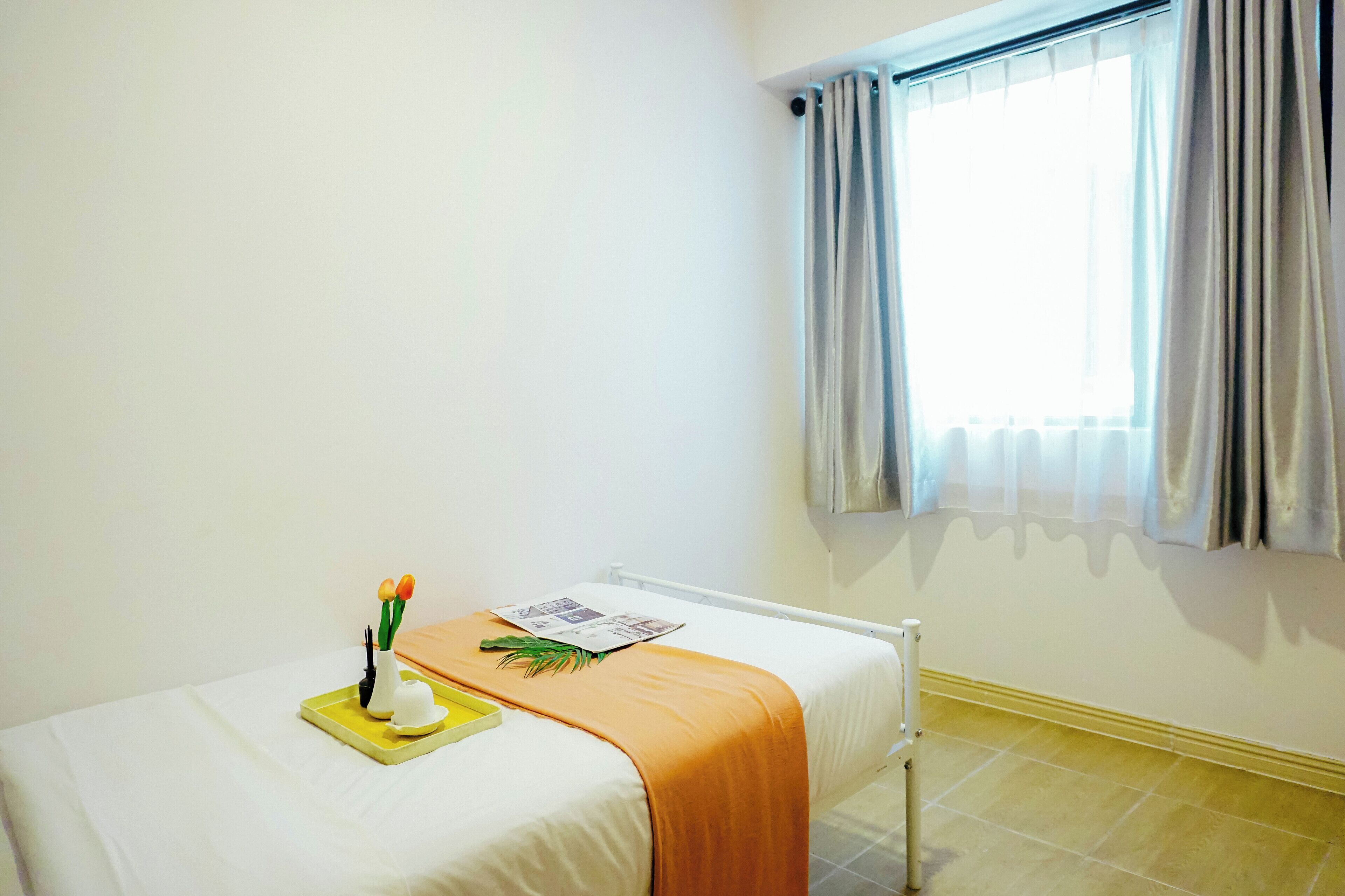 A hotel room with a bed, a window with curtains, a bedside table with a tray, and a small plant.