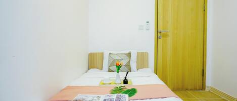 A neatly made bed with a magazine, a vase with flowers, and a small table with a cup and a phone charger.