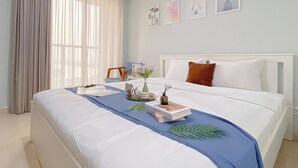 Un lit bien fait, recouvert d’une couvre-lit bleue, avec une tête de lit blanche et un oreiller blanc. Des tableaux encadrés ornent le mur, ainsi qu’une chaise et une fenêtre agrémentée de rideaux légers.