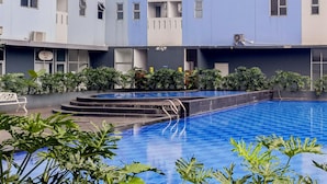A swimming pool with steps leading into it, surrounded by greenery and a multi-story building with balconies.