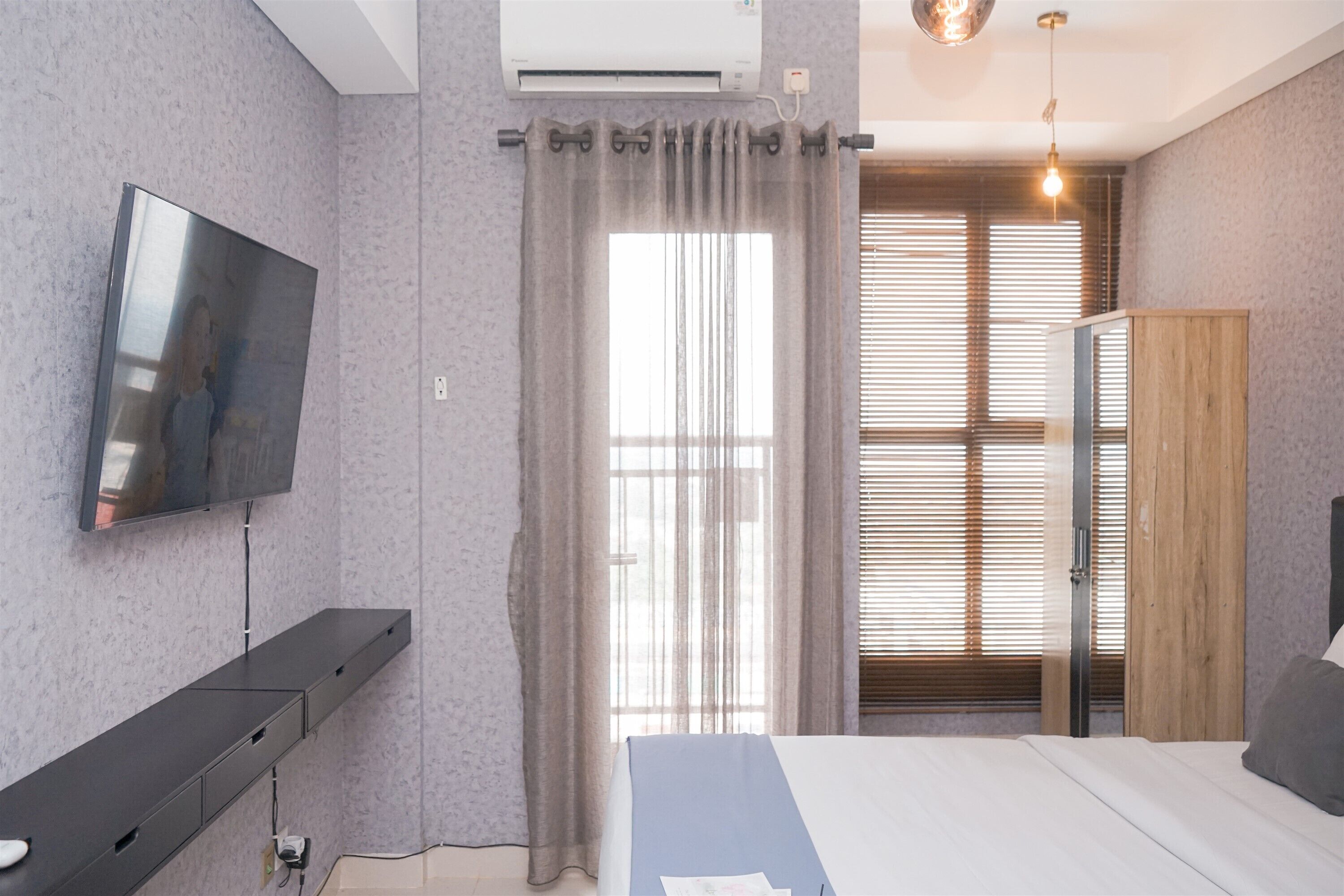 A modern bedroom with a flat-screen TV, a bed with a blue and white bedspread, a wooden wardrobe, and a window with blinds.