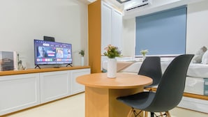 A modern hotel room with a TV on the wall, a round wooden table, black chairs, a white vase with flowers, and a large window with a view. - Cozy Living Studio Room At Meikarta Apartment (Cikarang)