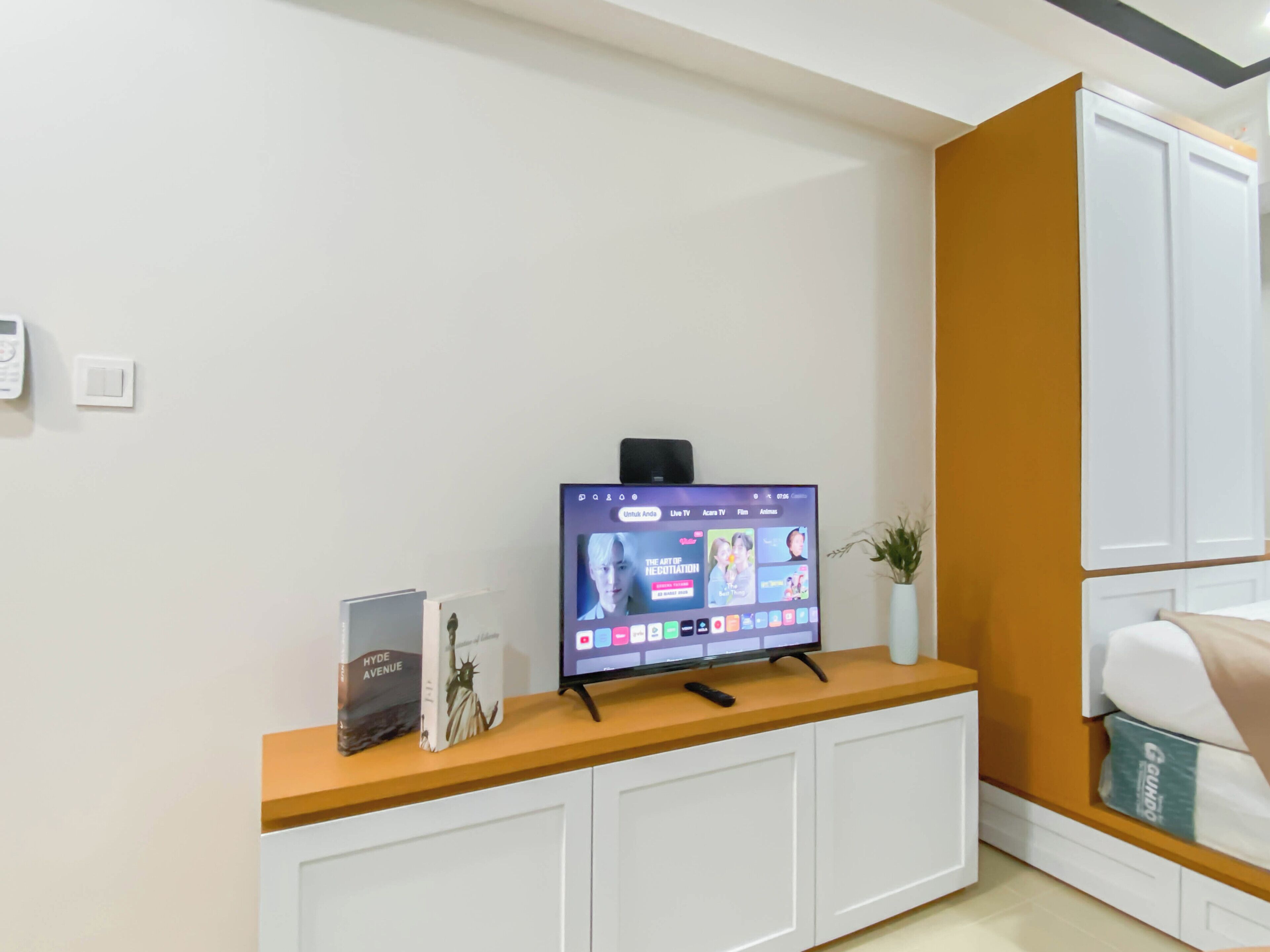 Habitación de hotel moderna con televisión de pantalla plana sobre una consola de madera, una planta pequeña en maceta y una cama con cabecera blanca.