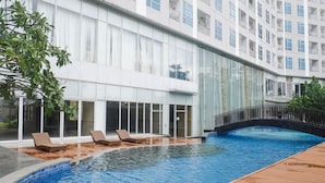 A swimming pool with lounge chairs, a wooden deck, and a building with large windows and balconies. - New Contemporary 2Br At Grand Sungkono Lagoon Apartment (Surabaya)