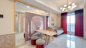 A modern living room with a large mirror, a wooden table, two red ottomans, a sofa, and a view of the city through large windows with pink curtains. - New Contemporary 2Br At Grand Sungkono Lagoon Apartment (Surabaya)