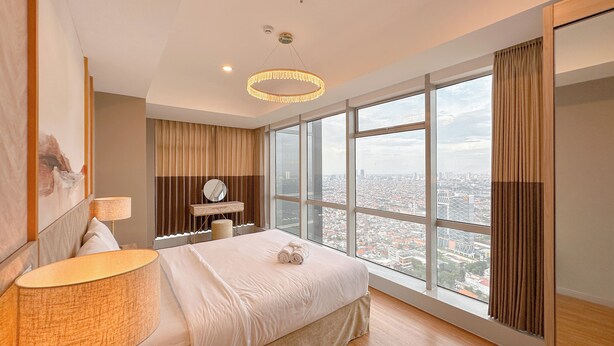 A modern bedroom with a large bed, a bedside lamp, a dresser, and a view of the city through large windows. - New Contemporary 2Br At Grand Sungkono Lagoon Apartment (Surabaya)