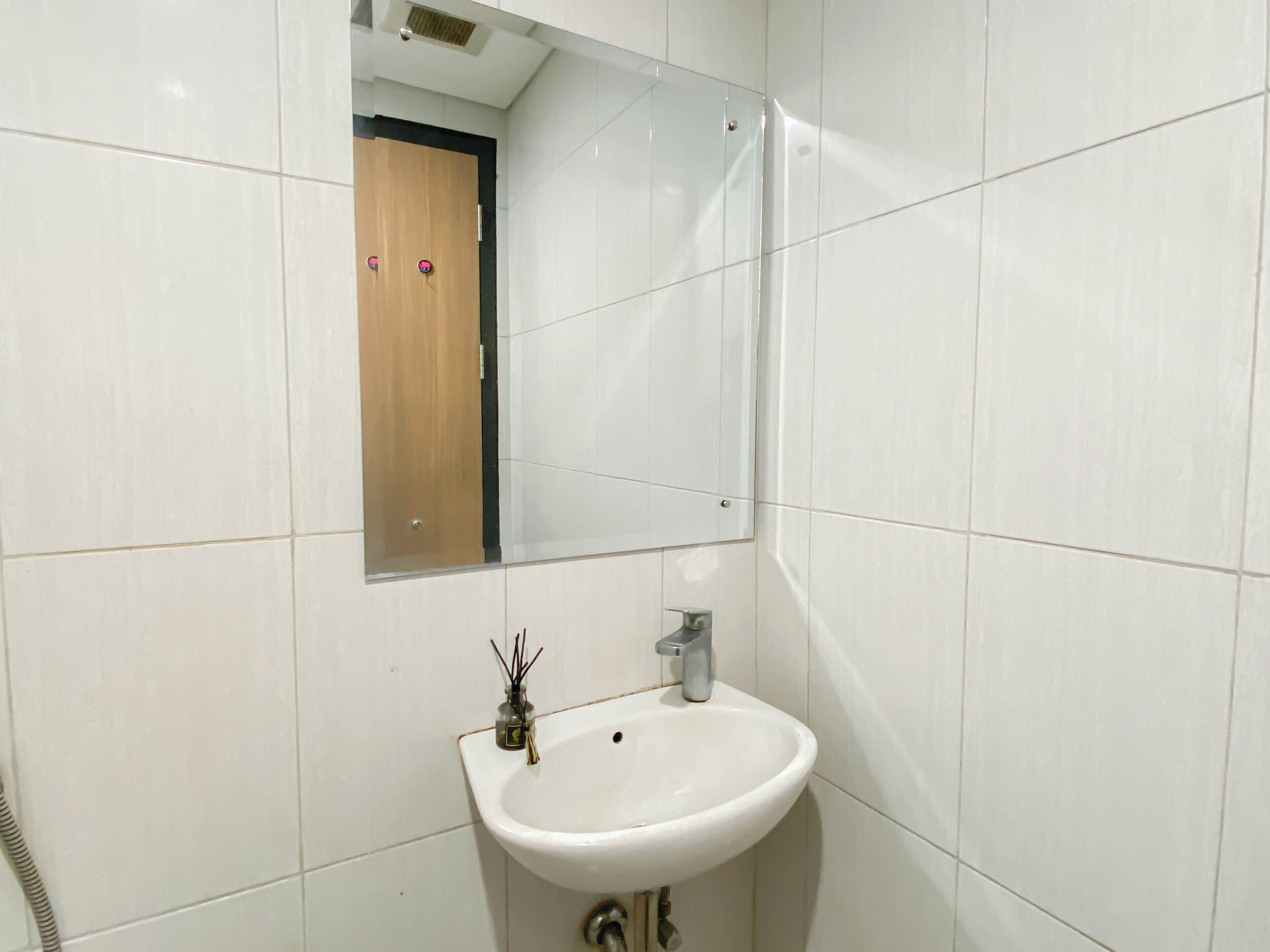 A bathroom with white tiles, a white sink with a single faucet, a mirror above the sink, and a wooden door in the background.