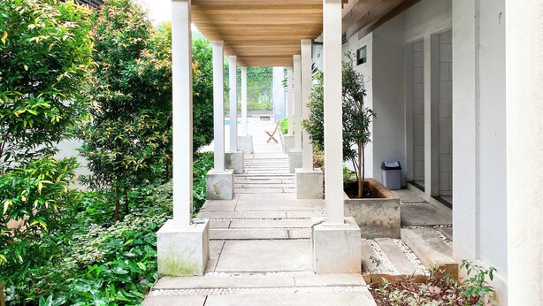 A covered walkway with columns, surrounded by greenery and leading to a pool area. - Tidy And Restful Studio At Monroe Tower Apartment (Cikarang)