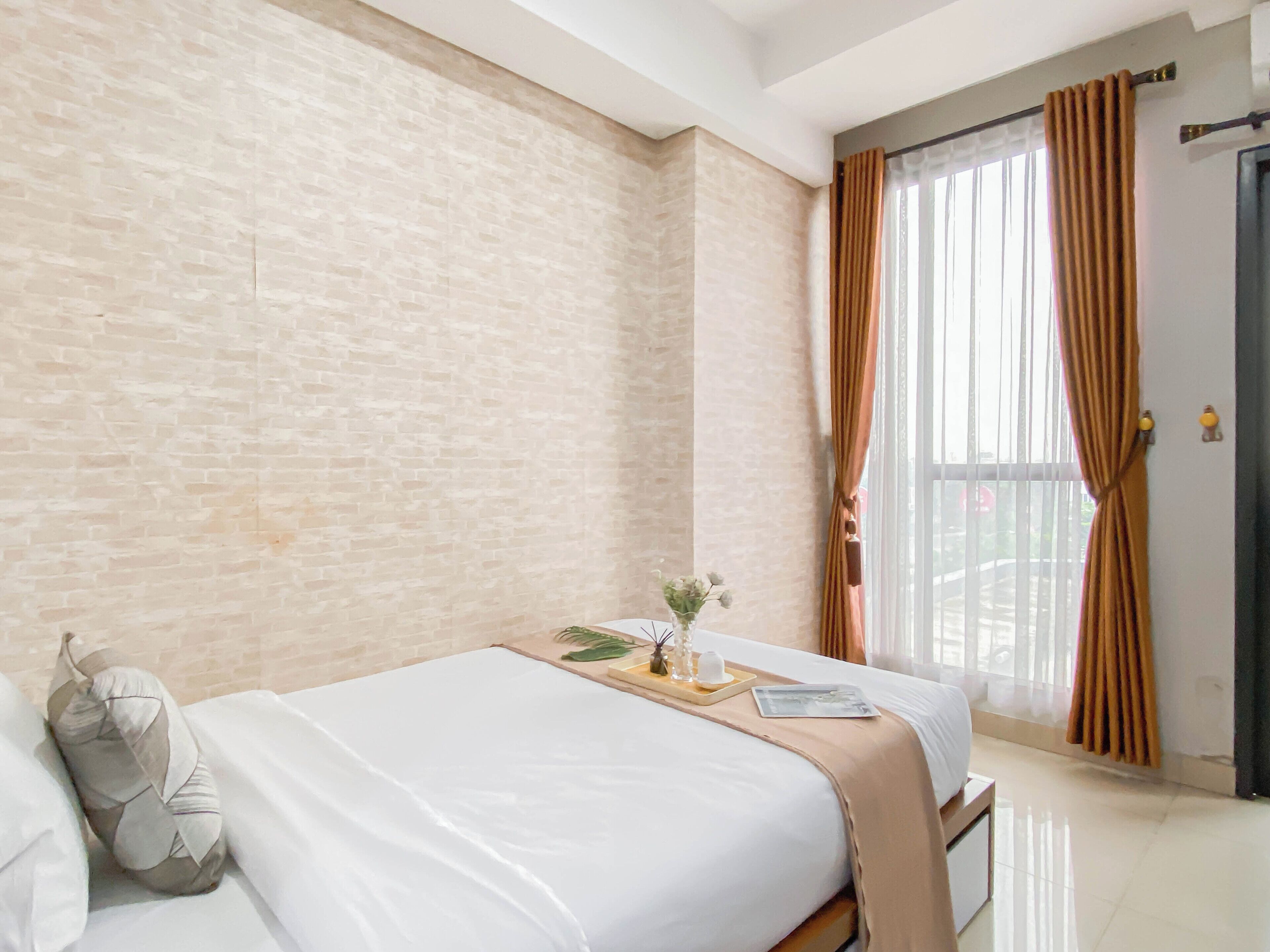 A bedroom with a brick wall, a bed with white bedding, a bedside table with a vase of flowers, and a window with curtains.