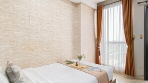 A bedroom with a brick wall, a bed with white bedding, a bedside table with a vase of flowers, and a window with curtains. - Tidy And Restful Studio At Monroe Tower Apartment (Cikarang)
