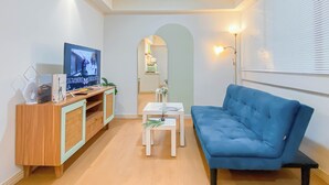Een moderne woonkamer met een blauwe bank, een houten tv-meubel, een klein wit tafeltje, en een keuken die zichtbaar is door een boog.