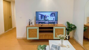 Een woonkamer met een televisie op een houten standaard, een tafeltje met een bloevas, en een deur op de achtergrond.