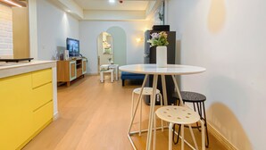 Een kleine keuken met een geel kastje, een witte tafel met krukken, en een televisie op een houten dressoir.
