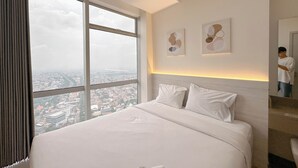 A hotel room with a large bed, a view of the city through a window, and two wall-mounted artworks. - Modern Suite 1Br At Grand Sungkono Lagoon Apartment (Surabaya)