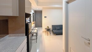 A modern kitchen with a built-in oven, a dining area with a small round table and two bar stools, and a grey sofa in a living area. - Modern Suite 1Br At Grand Sungkono Lagoon Apartment (Surabaya)