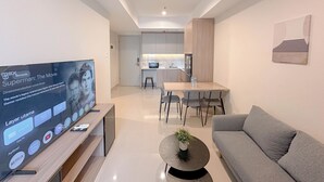 A modern living room with a flat-screen TV, a grey sofa, a coffee table with a plant, and a dining area with a table and chairs. - Stunning And Simply 2Br At Grand Sungkono Lagoon Apartment (Surabaya)
