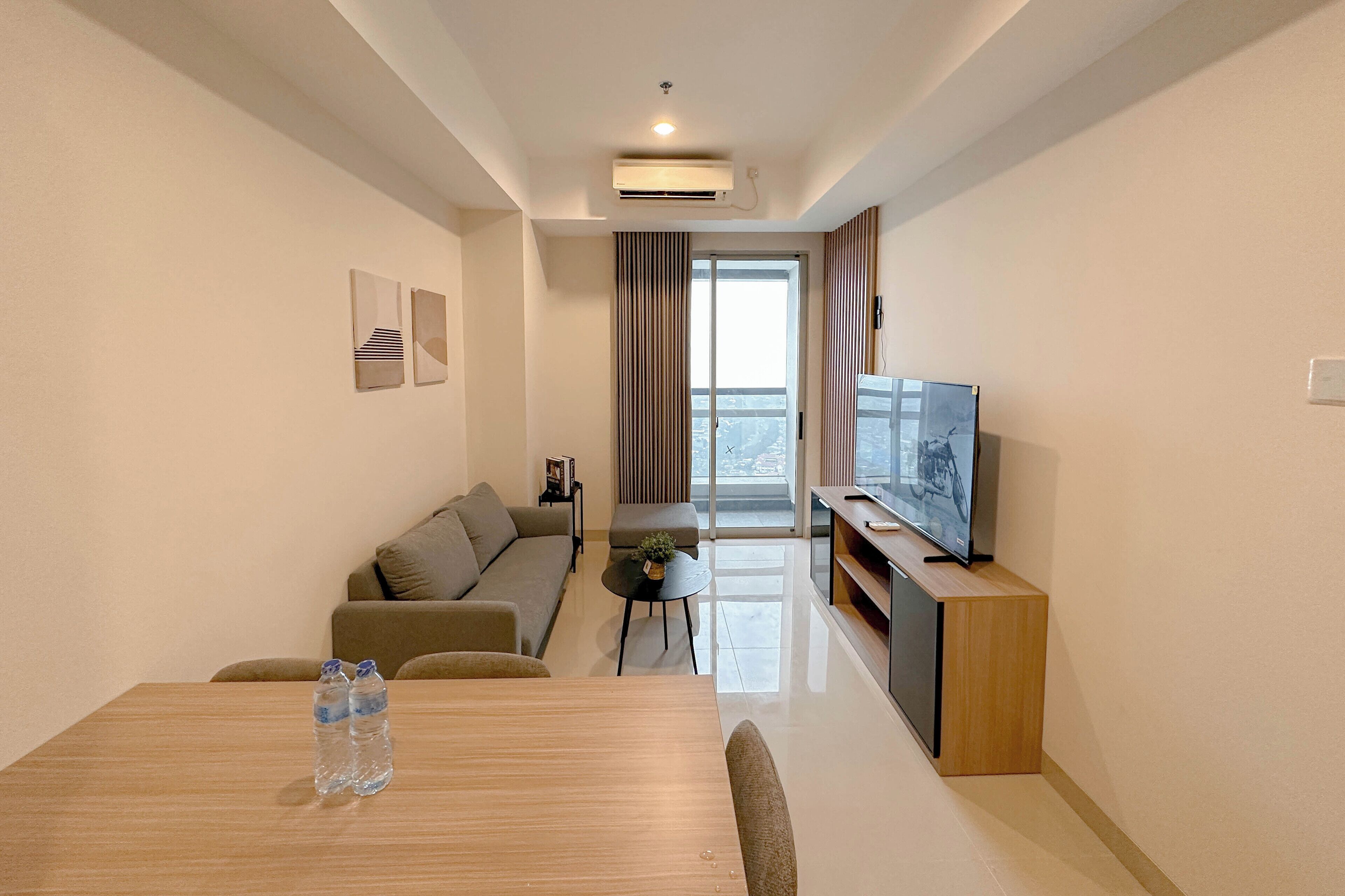 A modern living room with a sofa, a dining table with water bottles, a TV on a wooden stand, and a large window with curtains.