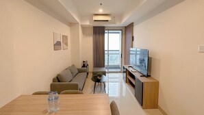 A modern living room with a sofa, a dining table with water bottles, a TV on a wooden stand, and a large window with curtains. - Stunning And Simply 2Br At Grand Sungkono Lagoon Apartment (Surabaya)