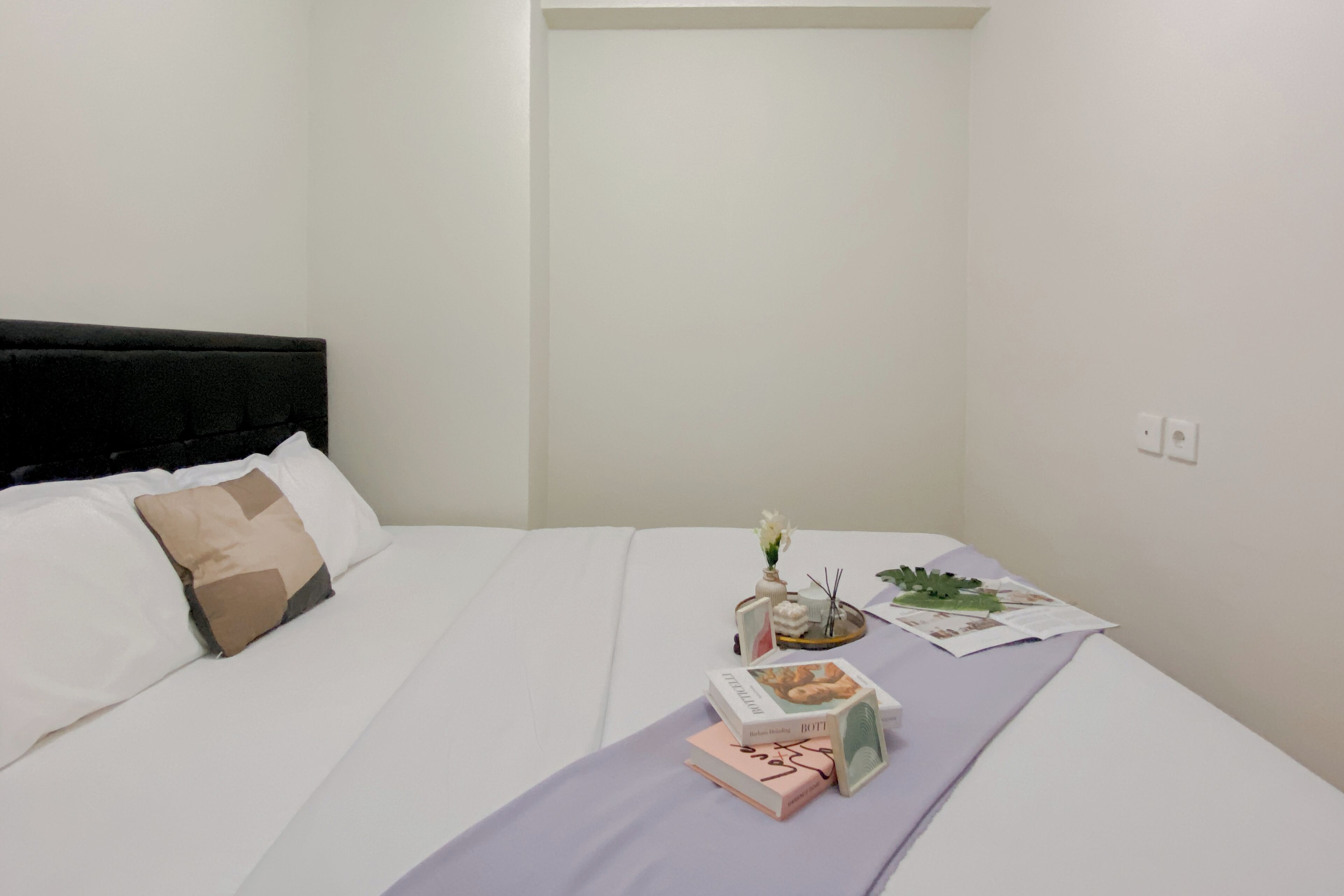 A neatly made bed with a white comforter and a lavender throw blanket. There is a small bedside table with a vase of flowers, a book, and a box. The room has white walls and a black headboard.