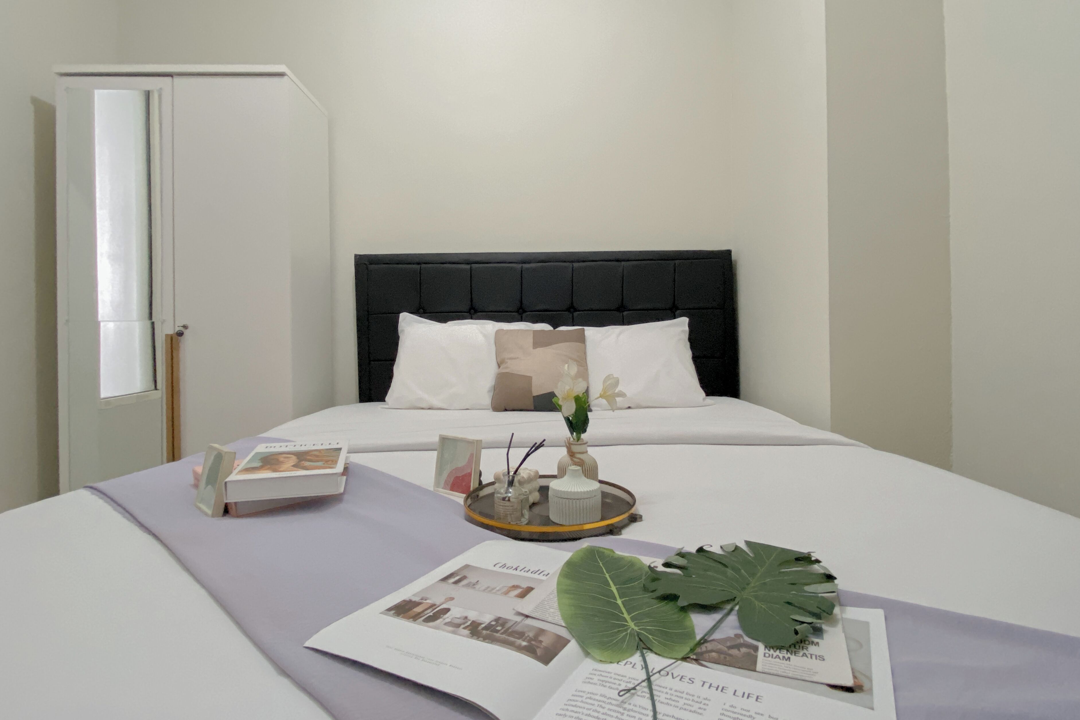 Een netjes opgemaakt bed met een zwart hoofdeinde, witte kussens en een dienblad met een boek, een vaas met bloemen en een blad.
