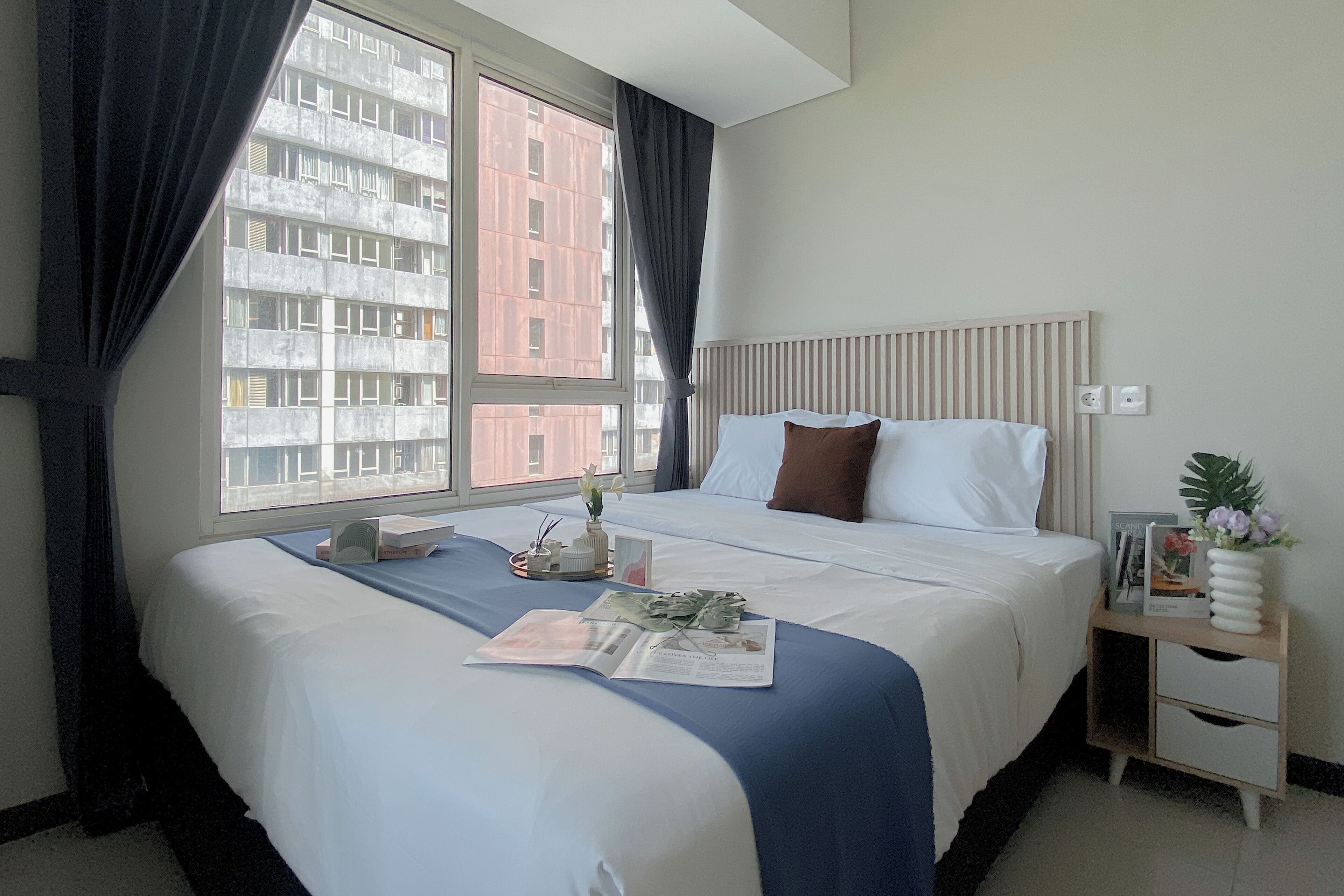 Una habitación de hotel con una cama grande, una mesita de noche con un florero, una ventana con cortinas y vistas a un edificio.