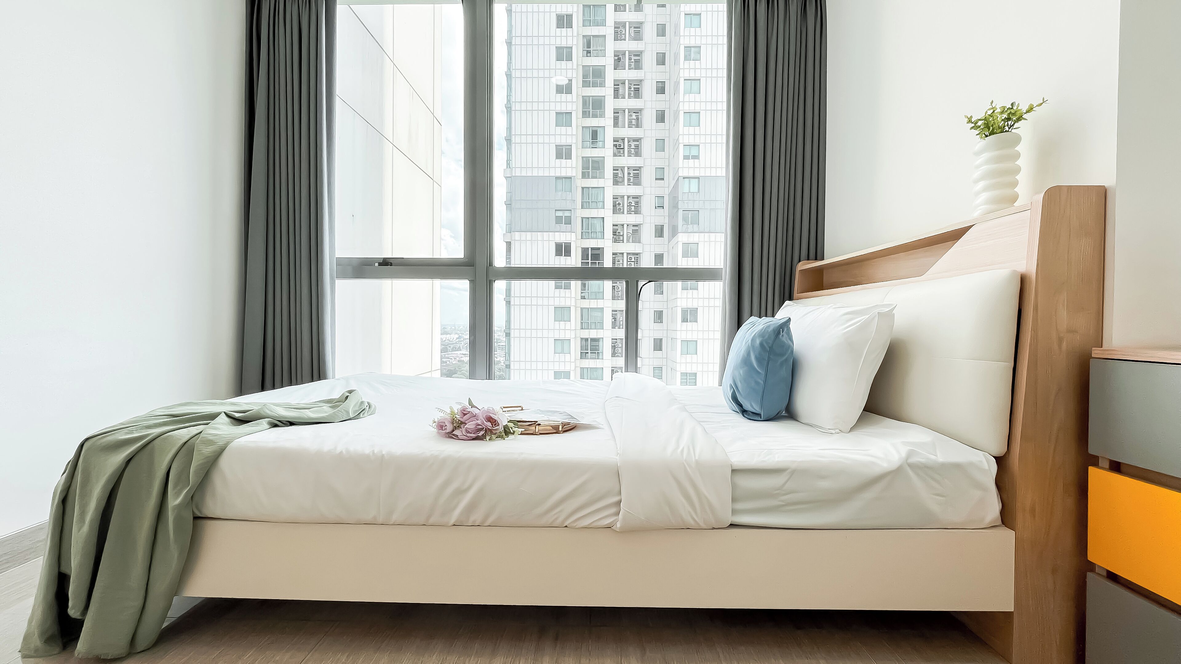 A modern bedroom with a large window, a bed with white bedding, a green throw, and a bedside plant.
