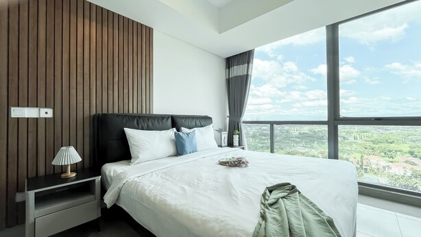 Una habitación de hotel moderna con una cama grande, una mesita de noche, una lámpara y vistas a un paisaje urbano a través de un amplio ventanal.