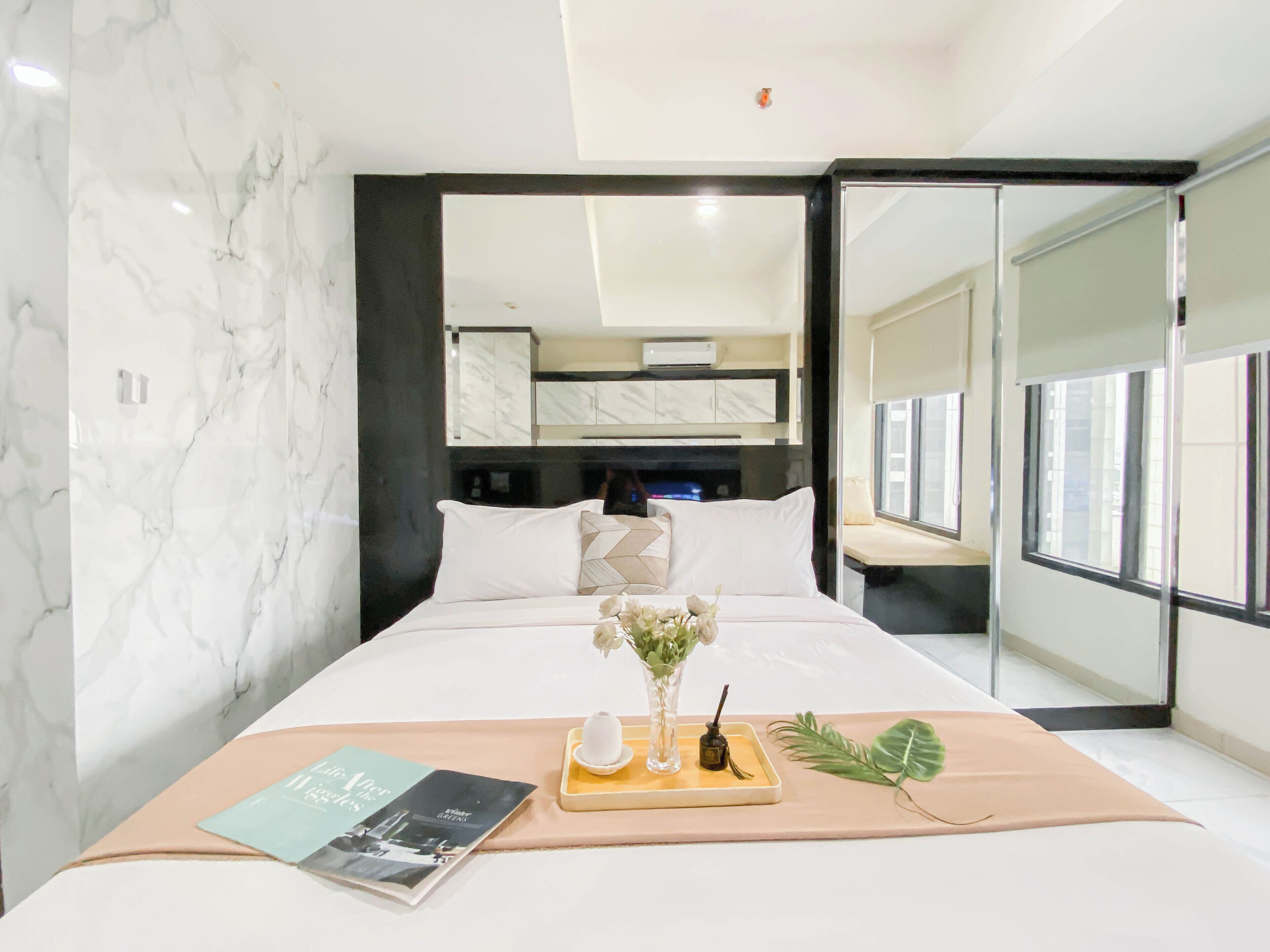 A modern hotel room with a marble wall, a bed with white bedding, a bedside table with a book and a vase of flowers, and a large window offering a view.
