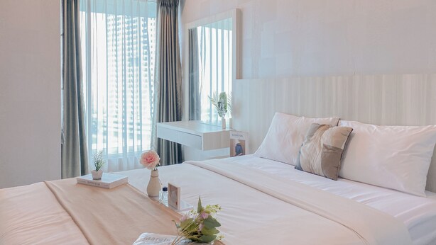 A hotel room with a large bed, a bedside table with a vase and a book, and a window with curtains. - Good Choice And Homey Studio Grand Kamala Lagoon Apartment (Bekasi)