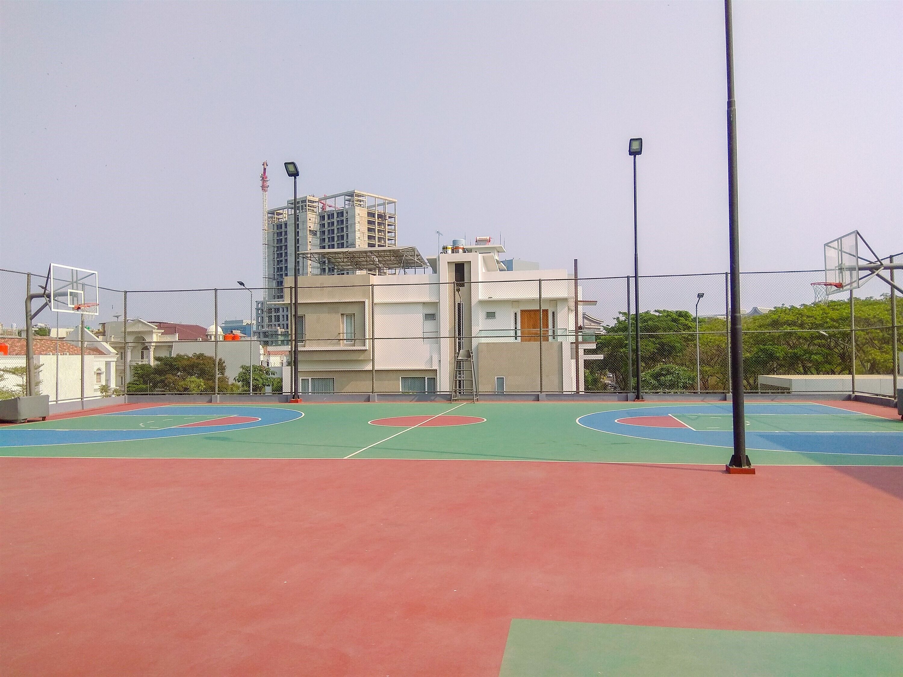 An outdoor basketball court with a red and green playing surface, surrounded by a metal fence.