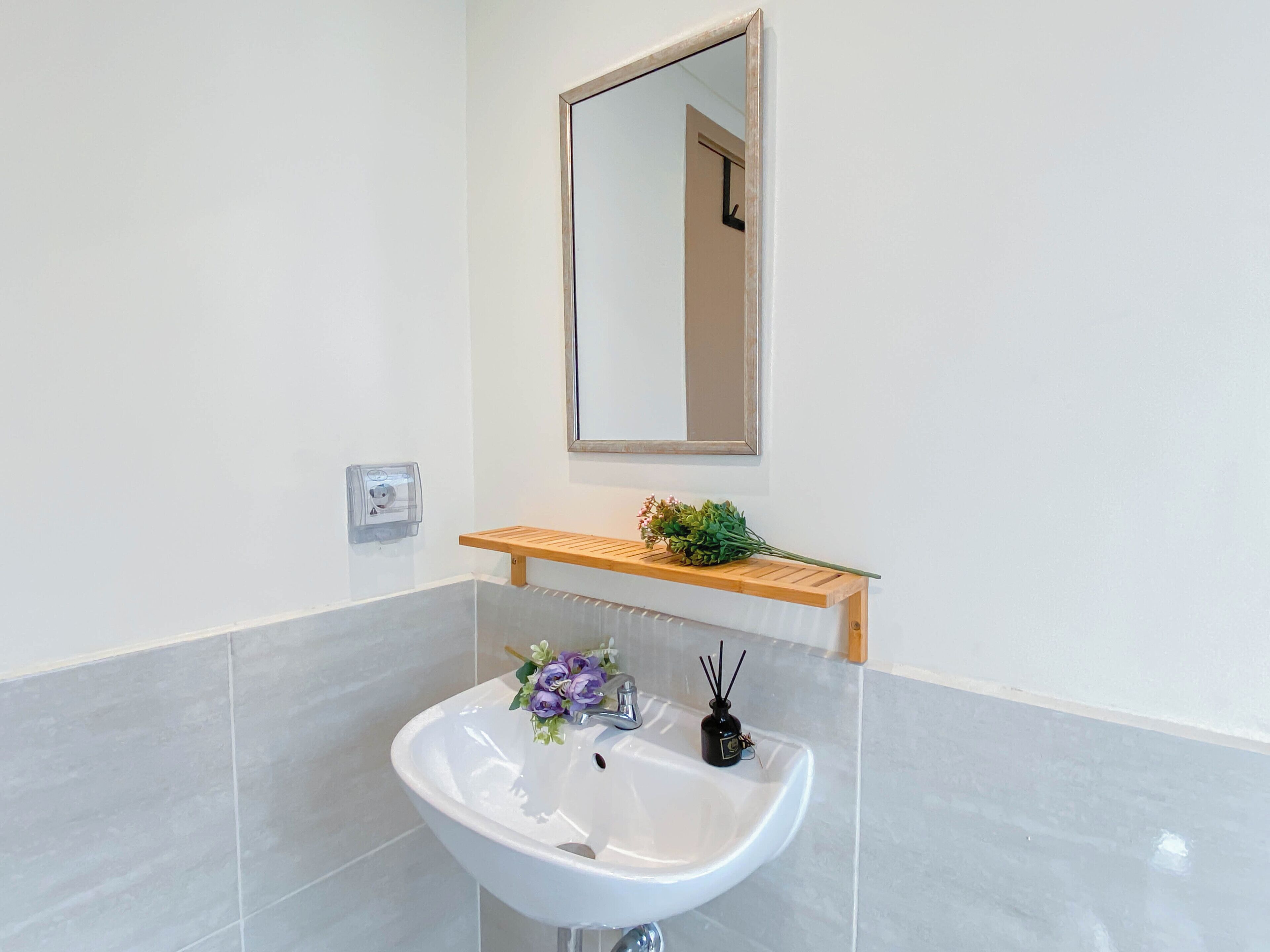 Ein Badezimmer mit einem weißen Waschbecken, einem Spiegel und einem Holzregal mit einer Pflanze.