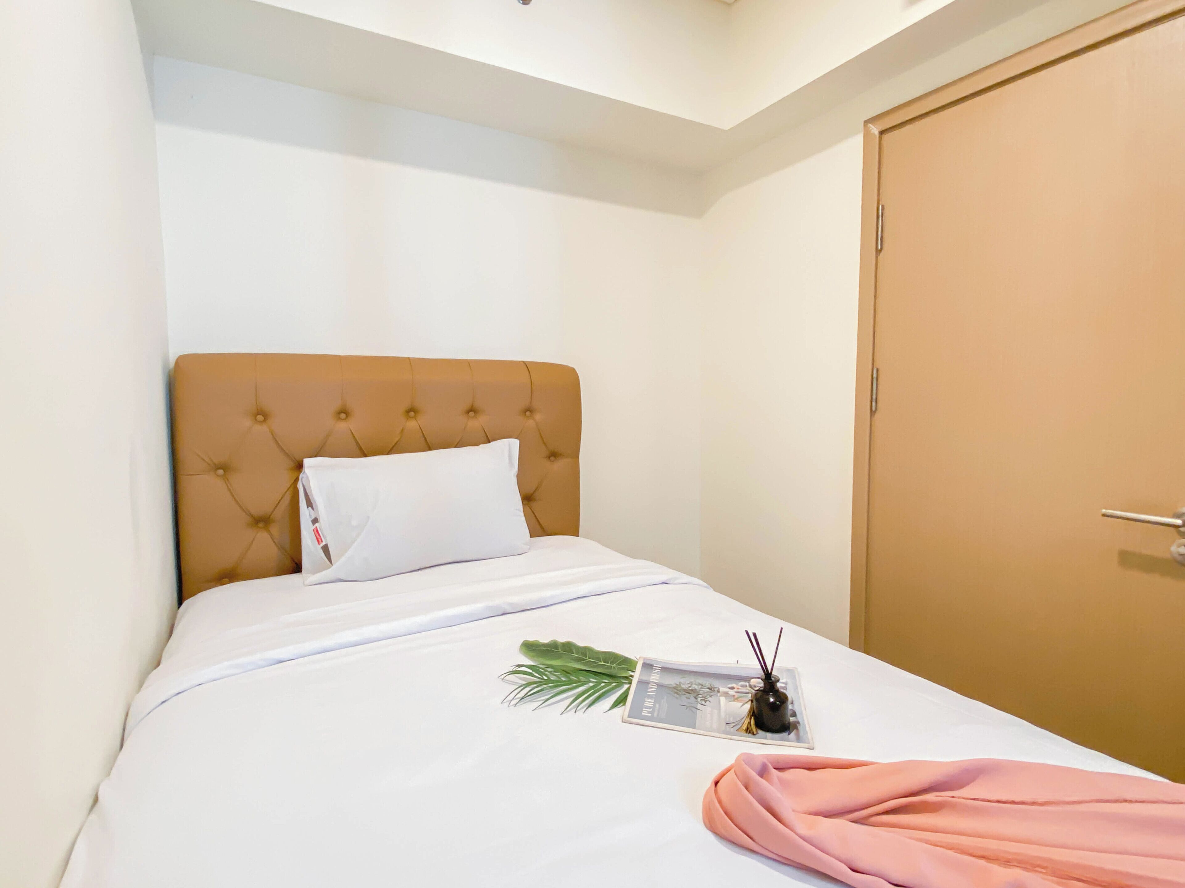A neatly made bed with a white comforter, a pink blanket, and a white pillow. There is a tray with a plant, a bottle, and a glass container on the bed.