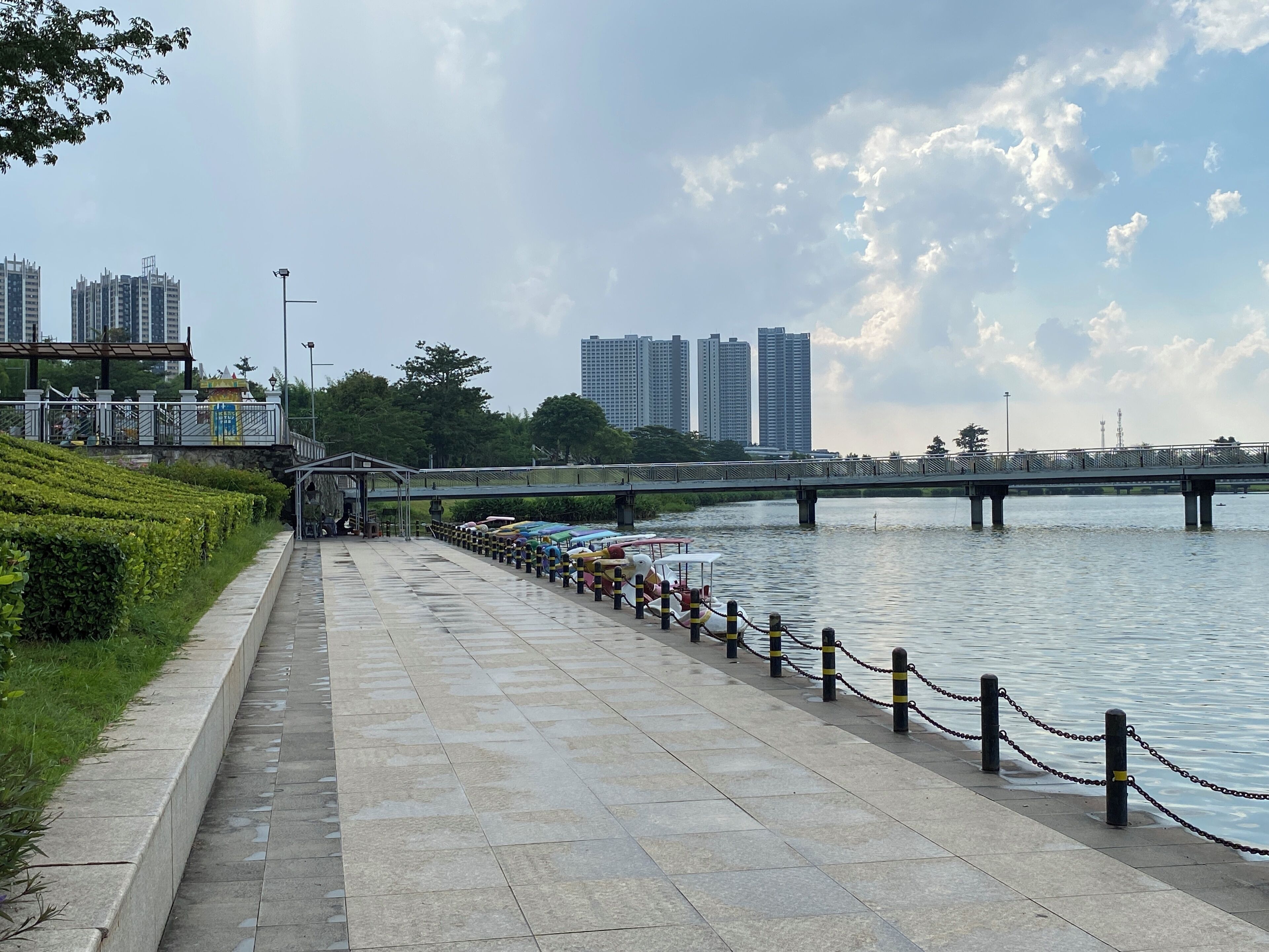 A riverside walkway with a bridge, benches, and a row of boats.